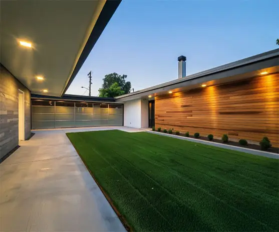 A sleek house with a green lawn, viewed from the left side, featuring a garage and concrete yard. Roof lights glow softly, enhancing the wooden accents on the walls.